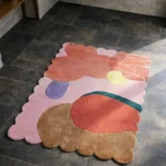 Perspective view of the Rose Riot rug placed on a dark tiled floor, showcasing the bold abstract color-block design and the structured pink scalloped border illuminated by natural sunlight.