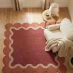 High angle view of the Rose Whisper rug on a wooden floor, highlighting the square shape and the playful contrast between the wavy border and scalloped edges.