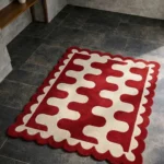 High angle view of the Ruby Weave rug placed on a dark tiled floor, featuring a bold interlocking wavy pattern in deep red and cream, finished with a decorative red scalloped edge.