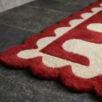 Close-up detail of the Ruby Weave rug, highlighting the plush hand-tufted wool texture, the distinctive cream and red wavy interlocking design, and the intricate deep red scalloped border.