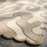 Close-up angled view of the Sand Drift rug illuminated by sunlight, highlighting the plush hand-tufted New Zealand wool texture, the deeply carved lines defining the abstract beige shapes, and the intricately contoured scalloped edge.