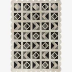 Top down view of the Shadow Grid rug against a white background, featuring a repeating patchwork pattern of geometric squares, triangles, and diamonds in shades of black, dark grey, light grey, and cream, framed by a continuous cream scalloped border.