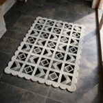 Perspective view of the Shadow Grid rug placed on a dark tiled floor, showcasing the bold monochromatic geometric patchwork design and the structured scalloped border illuminated by natural sunlight.