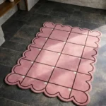 High angle view of the Soft Depth rug featuring a soft pink background with a structured dark brown grid pattern, small cream square accents at the intersections, and a prominent scalloped border.