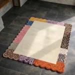 Perspective view of the Soft Frame rug placed on a dark tiled floor, featuring a solid cream center surrounded by a vibrant, multi-colored scalloped border with playful embossed squiggly lines.