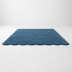 Low angle view of the Still Tides rug lying flat against a white background, featuring a solid deep blue color with horizontally carved ribbed lines and a continuous scalloped edge.