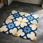 Perspective view of the Sun Dot Posy square rug placed on a dark tiled floor, featuring a vibrant retro floral pattern with large cream flowers, mustard yellow centers, and smaller light blue accents on a deep navy blue background, finished with a petal-contoured scalloped edge.