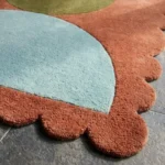 Close-up angled view of the Sunbaked Form rug, highlighting the dense hand-tufted New Zealand wool and viscose silk texture, the deeply carved lines separating the terracotta, blue, and green geometric shapes, and the intricately contoured scalloped edge.