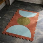 Perspective view of the Sunbaked Form rug placed on a dark tiled floor, showcasing the striking geometric design and the structured terracotta scalloped border illuminated by natural sunlight.