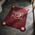 High angle view of the Velvet Bloom rug placed on a dark tiled floor, featuring a deep burgundy background with a contrasting light pink line-art rose illustration and unique rounded corners containing pink circles.