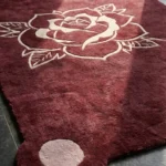 Close-up detail of the Velvet Bloom rug, highlighting the plush hand-tufted wool texture, the embossed pink line-art of a blooming rose, and the distinctive rounded corner with a solid pink accent circle.