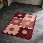 High angle view of the Velvet Garden rug placed on a dark tiled floor, featuring a retro large-scale floral pattern in shades of soft pink and deep burgundy with tan centers, finished with a unique scalloped edge that contours to the shape of the petals.