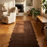 Perspective lifestyle view of the Walnut Crest runner rug placed on a warm wooden floor in a sunlit living room with a fireplace, featuring a solid dark brown color and a continuous contoured scalloped edge.