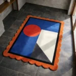 High angle view of the Amber Solis rug featuring a geometric design with a bold orange sun circle on a deep blue background, accented by white and grey angular shapes and a rust scalloped border.