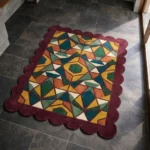 High angle view of the Autumn Prism rug featuring an intricate kaleidoscopic pattern with teal, mustard, and orange shapes framed by a deep burgundy scalloped border.