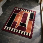 High angle view of the Burnt Frame rug featuring a modern geometric design with interlocking blocks in rust, burgundy, and black, surrounded by a distinctive black and cream striped inner border and a deep wine scalloped edge.