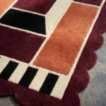 Close-up detail of the Burnt Frame rug's corner, showcasing the plush hand-tufted wool texture, the geometric black and cream dashed border, and the deep burgundy scalloped edge.