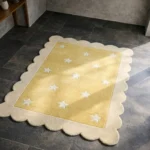 Perspective view of the Buttercream Bloom rug placed on a dark tiled floor, highlighting the soft yellow center adorned with scattered white stars and framed by a cream scalloped border.