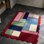 Perspective view of the Canvas Mosaic rug placed on a dark tiled floor, highlighting the vibrant geometric color-block pattern in pink, blue, green, and cream, framed by a red scalloped border.