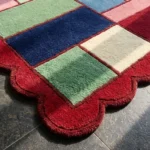 Close-up detail of the Canvas Mosaic rug's corner, showcasing the plush hand-tufted wool texture, the colorful geometric blocks, and the intricately carved red scalloped edge.