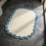 Perspective view of the Cloud Marina rug placed on a dark tiled floor, highlighting the airy cream tones and the playful blue scalloped border.