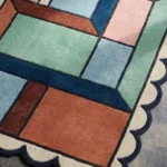 Close-up detail of the Dusty Dune rug's corner, showcasing the thick wool pile, precise color carving, and the decorative blue scalloped edge.