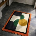 High angle view of the Everne Meadow rug featuring a modern geometric design with green and mustard yellow curved shapes on a cream and black background, framed by a vibrant orange scalloped border.