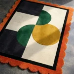 Close-up perspective of the Everne Meadow rug, highlighting the plush hand-tufted wool texture and the striking contrast of the orange scalloped edge against the black and cream geometric pattern.