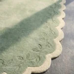 Close-up detail of the Garden Lace rug's corner, showcasing the plush hand-tufted wool texture and the embossed leafy vine motif on the cream scalloped edge.