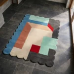 High angle view of the Gentle Pulse rug featuring a modern abstract design with interlocking shapes in light blue, rust, and cream, accented by a bright red square and framed by a dark grey scalloped border.