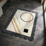 High angle view of the Incorix rug featuring a minimalist geometric design with diagonal lines and bold black shapes on a cream background, framed by a dark grey scalloped border.