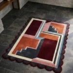High angle view of the Inverse Staircase rug featuring an architectural 3D design with geometric steps in rust, burgundy, pink, and dark grey, framed by a deep red scalloped border.
