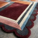 Close-up detail of the Inverse Staircase rug's corner, highlighting the plush hand-tufted wool texture, the crisp layered geometric lines in pink, cream, and light blue, and the intricate deep burgundy scalloped edge.