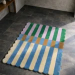 Perspective view of the Mint Gelato rug placed on a dark tiled floor, showcasing the bold, segmented striped design and the contoured scalloped edge illuminated by natural sunlight.