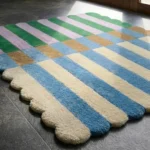 Close-up angled view of the Mint Gelato rug near a sunlit doorway, highlighting the plush hand-tufted New Zealand wool and viscose silk texture, the clean lines separating the vibrant color-block stripes, and the intricately sculpted scalloped edge.