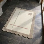 High angle view of the Mist Alba rug featuring a minimalist design with delicate sage green and dusty pink lines on a cream background, framed by a beige scalloped border.