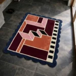 High angle view of the Muted Garnet rug featuring a modern geometric design with interlocking shapes in burgundy, rust, and pink, accented by a black and white striped pattern and framed by a deep blue scalloped border.