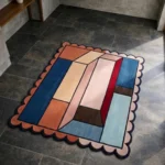 High angle view of the Muted Studio Square rug featuring a modern geometric color-block design with interlocking 3D-style shapes in blue, rust, and pink, framed by a blush scalloped border with dark trim.