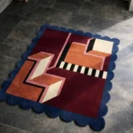 High angle view of the Noir Blocks rug featuring a modern geometric design with burgundy, rust, and cream interlocking shapes, accented by a black and white striped pattern and framed by a deep blue scalloped border.