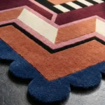 Close-up detail of the Noir Blocks rug's corner, highlighting the plush hand-tufted wool texture, the sharp geometric lines in rust and cream, and the contrasting blue scalloped edge.