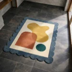 High angle view of the Ochre Loom rug featuring abstract organic shapes in mustard yellow, rust, and teal on a cream background, framed by a blue scalloped border.