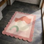 High angle view of the Peach Wisp rug featuring abstract organic shapes in soft peach, cream, and sage green, framed by a pink scalloped border.