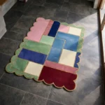 Perspective view of the Piatched Poise rug placed on a dark tiled floor, highlighting the vibrant geometric color-block pattern in pink, green, blue, and deep maroon, framed by a scalloped edge with a contrasting gold trim.