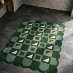 Perspective view of the Pine Patchwork rug placed on a dark tiled floor, showcasing the structured geometric checkerboard design and the wavy green scalloped border illuminated by natural sunlight.