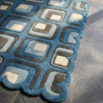 Close-up detail of the Retrose Orchid rug, showcasing the dense hand-tufted wool texture, the deeply carved and embossed lines separating the blue and grey geometric blocks, and the intricate scalloped border.
