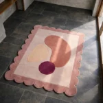 High angle view of the Rose Veil rug featuring abstract organic shapes in rose, cream, and deep purple on a blush background, framed by a dusty rose scalloped border.