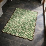 Perspective view of the Sage Dream rug placed on a dark tiled floor, showcasing the structured geometric floral trellis design and the contoured brown scalloped border illuminated by natural sunlight.