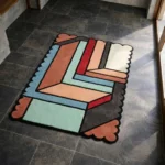 High angle view of the Sage Echo rug featuring a modern geometric color-block design with interlocking shapes in sage green, rust, and blue, defined by bold black outlines and a scalloped edge.