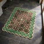 Perspective view of the Scroll Halo rug placed on a dark tiled floor, showcasing the structured geometric lattice, the decorative green tile border, and the contoured cream scalloped edge illuminated by natural sunlight.