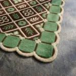 Close-up angled view of the Scroll Halo rug, highlighting the dense hand-tufted New Zealand wool and viscose silk texture, the deeply carved lines defining the lattice pattern, floral motifs, and green border tiles, along with the intricately contoured cream scalloped edge.